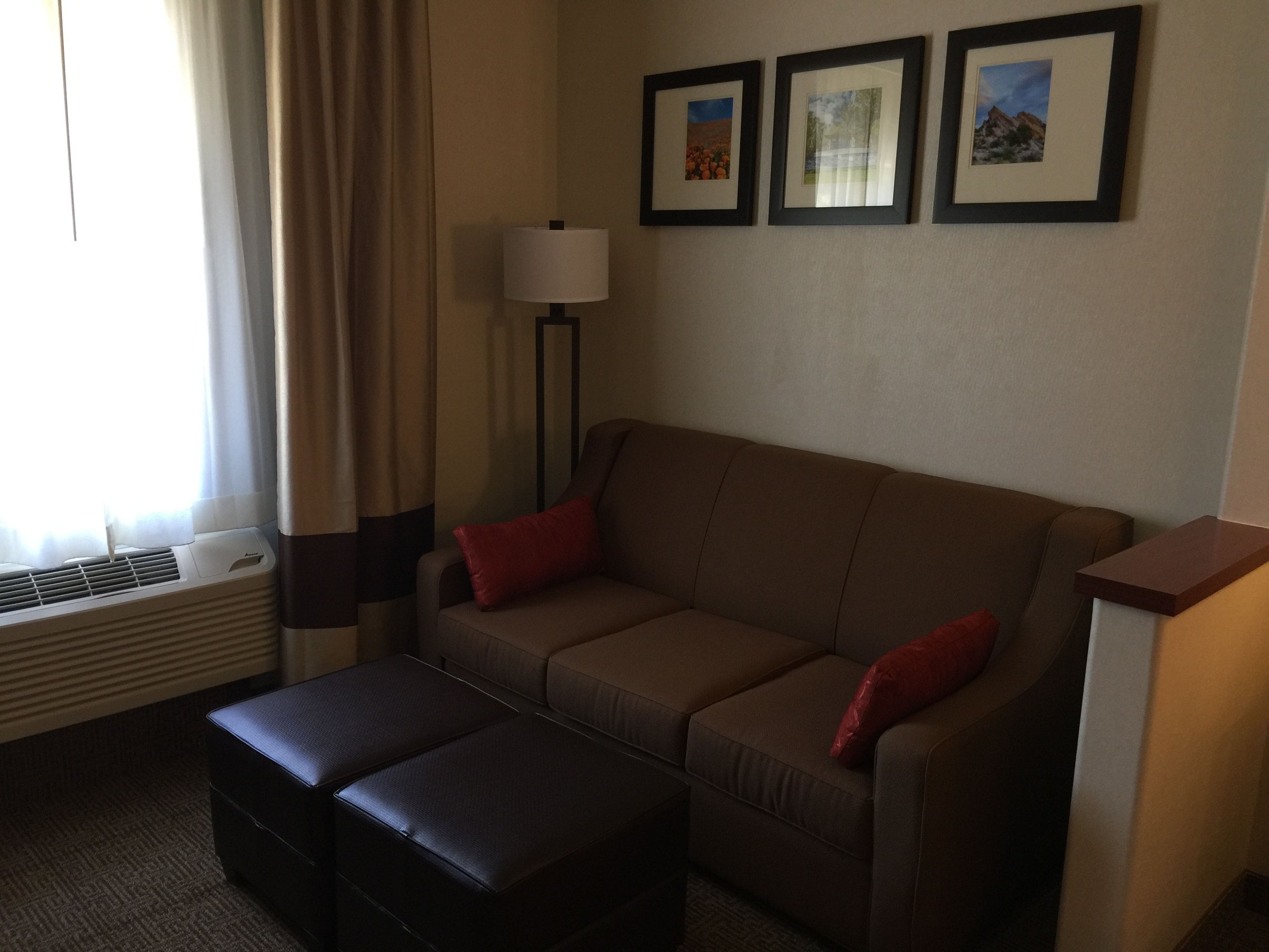Habitación de hotel con sofá marrón, dos cojines rojos, un otoman negro, una lámpara de pie y tres cuadros en la pared.
