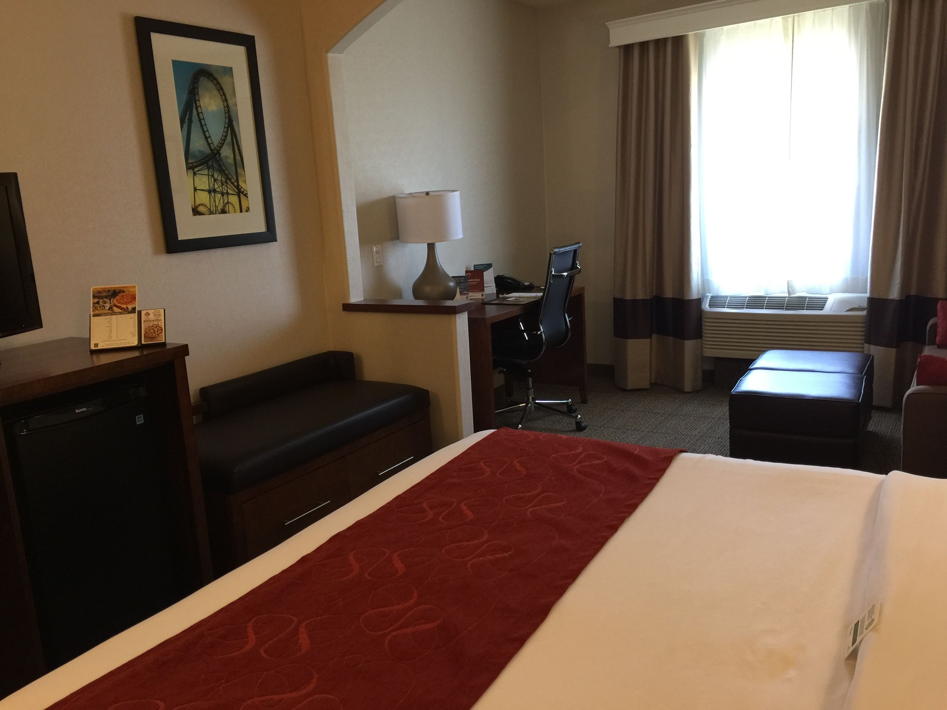 Habitación de hotel con una cama, un escritorio con silla, una mesita con lámpara, televisor y un cuadro en la pared.
