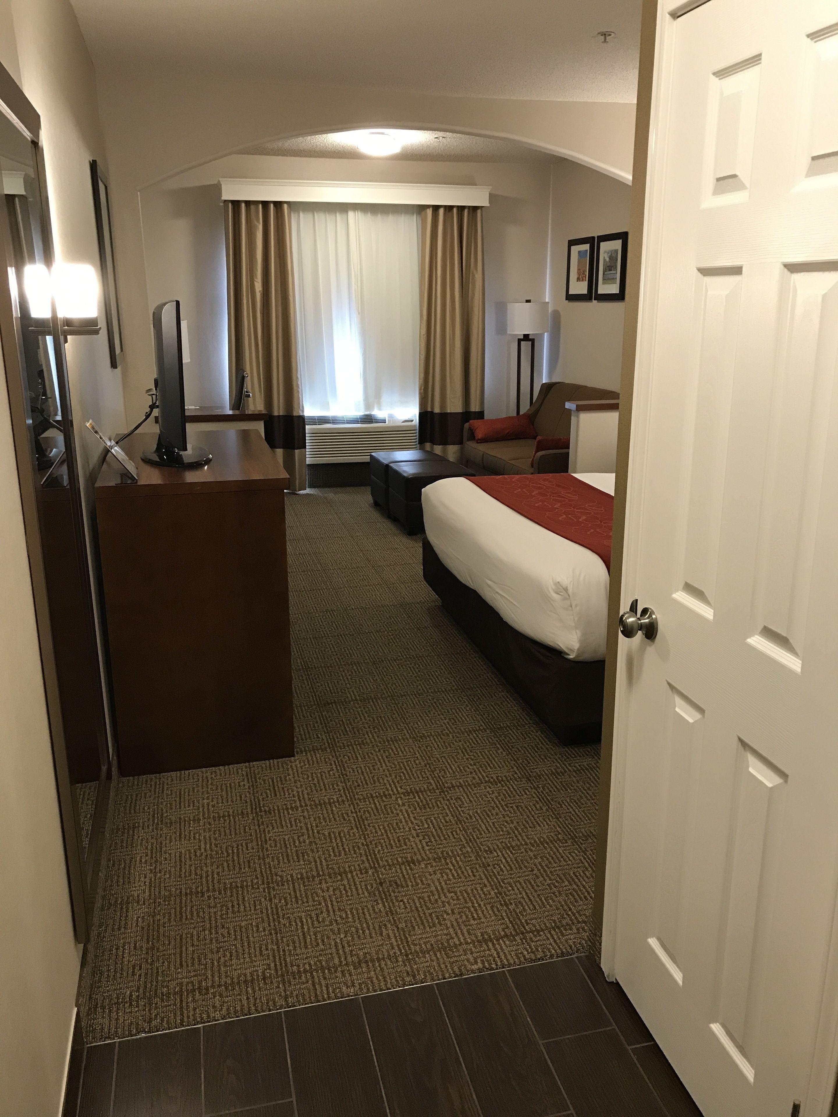 Une chambre d’hôtel avec un lit, un bureau avec un ordinateur, un canapé, une lampe et une fenêtre avec des rideaux.