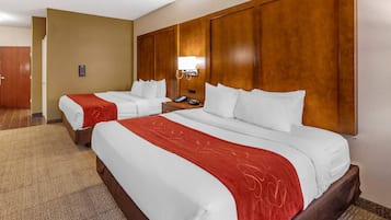 A hotel room with two beds, a wooden headboard, a bedside table with a lamp, and a carpeted floor.