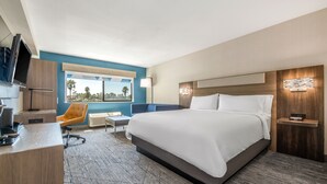 Una habitaci ón de hotel moderna con una cama grande, un escritorio con una computadora, una silla, un televisor y una ventana con vista a palmeras.