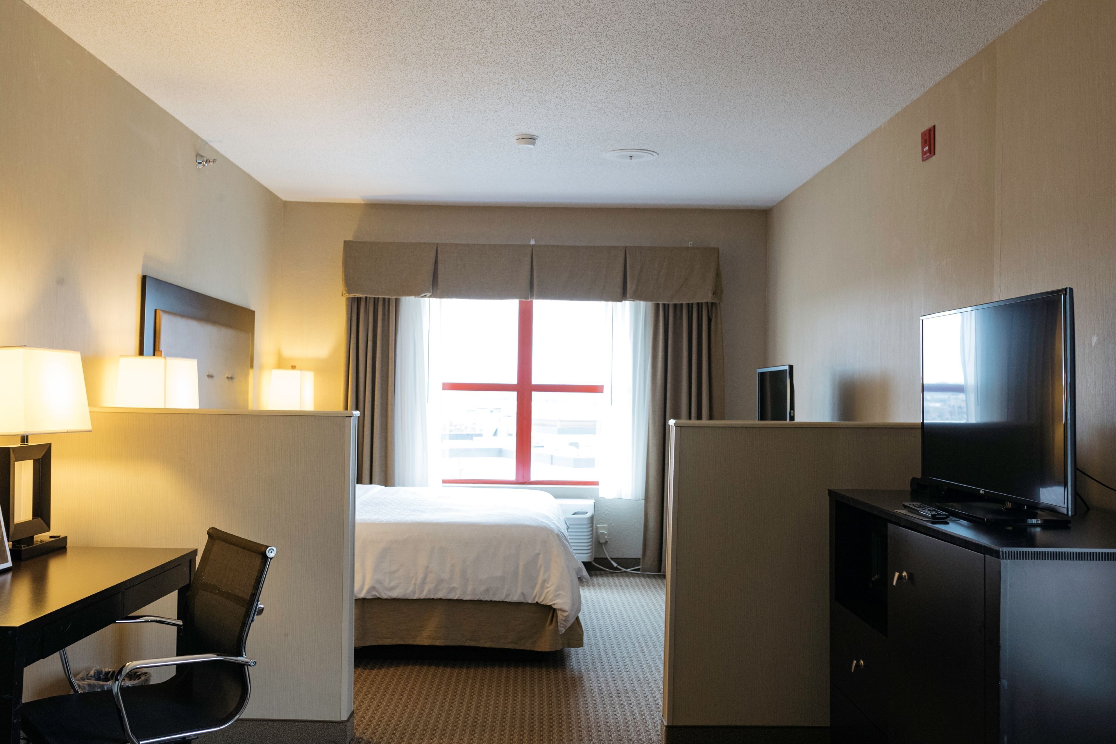 Una habitación de hotel con una cama, un escritorio con una silla, un televisor y un ventanal con cortinas.