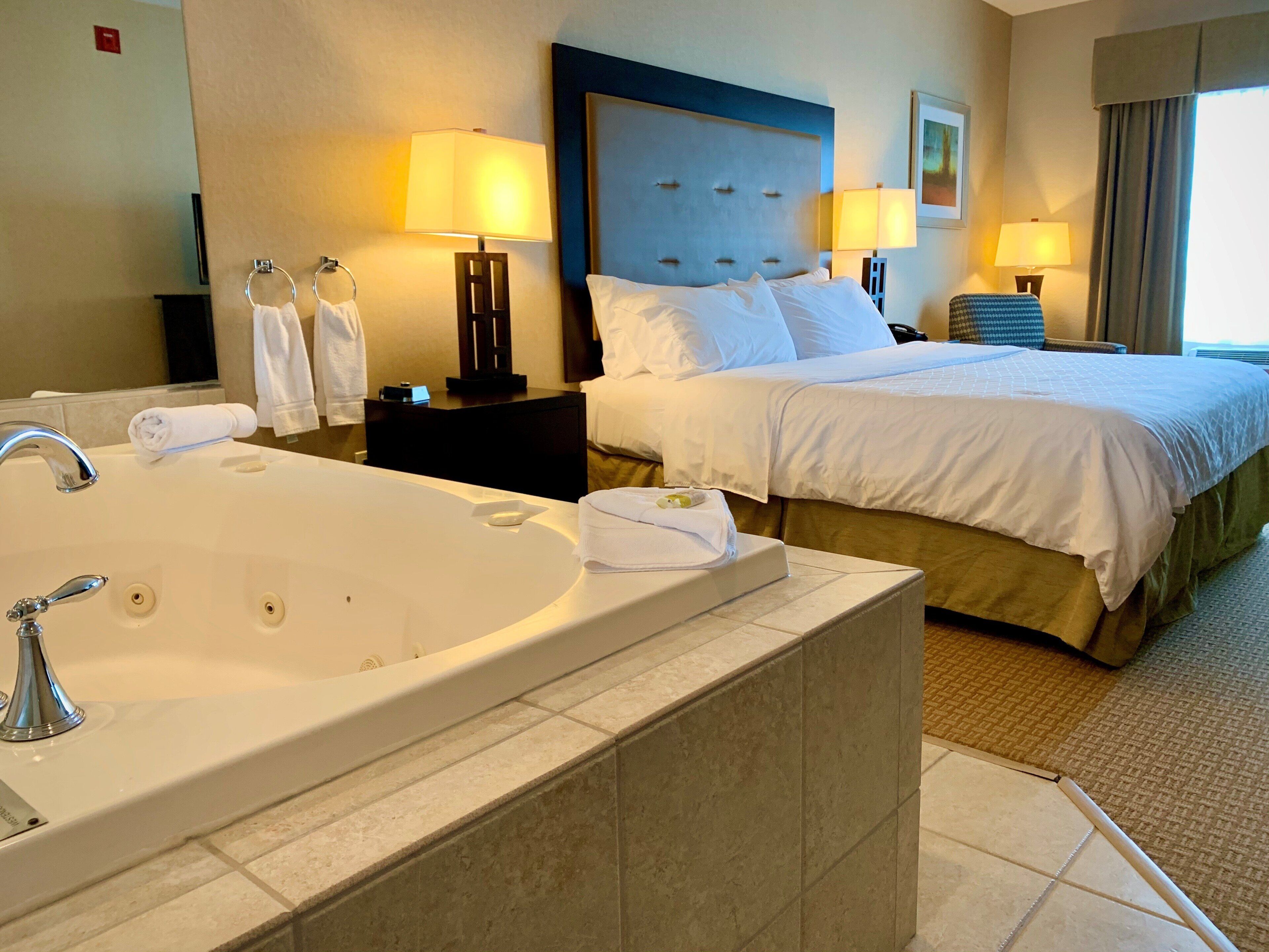 Habitación de hotel con una bañera grande, una cama con ropa blanca, dos lámparas de noche y un cabecero.