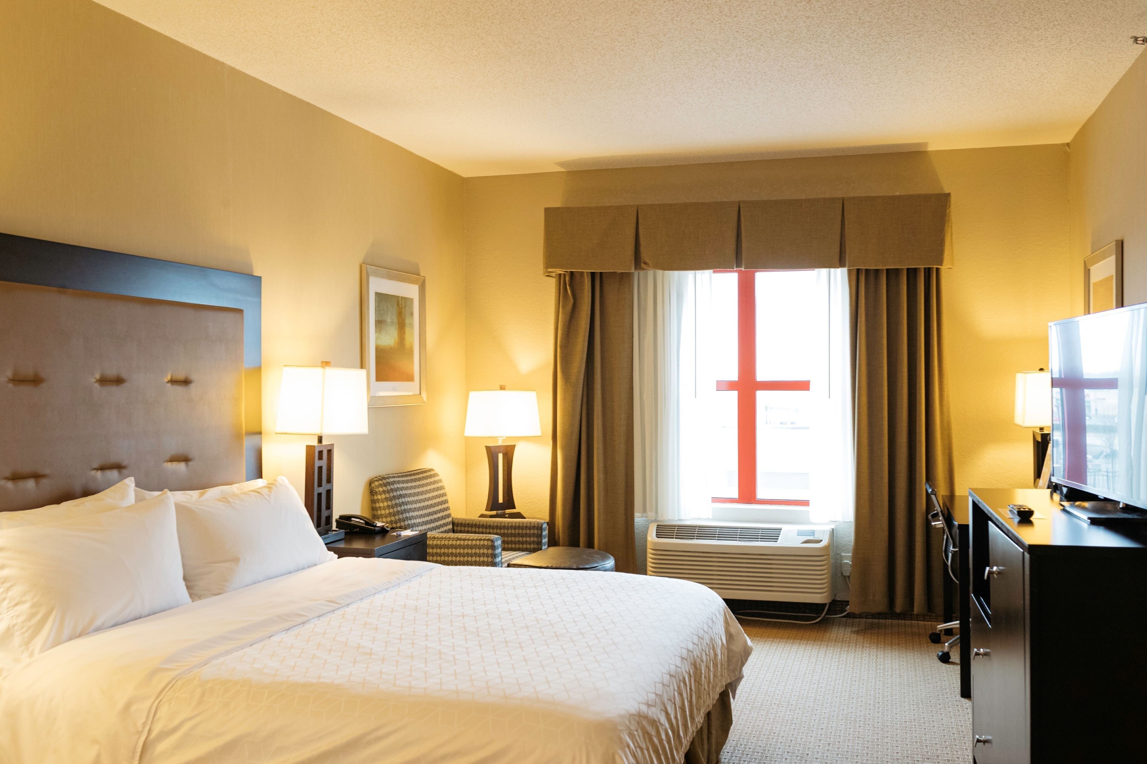 A hotel room with a large bed, a desk, a chair, a television, and an air conditioning unit.