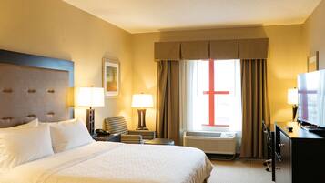 A hotel room with a large bed, a desk, a chair, a television, and an air conditioning unit.