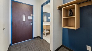 A hotel hallway with a wooden door, a blue-framed mirror, a wooden shelf with a decorative item, and a bathroom visible through an open door. - Spark by Hilton Houston near the Galleria (Houston)