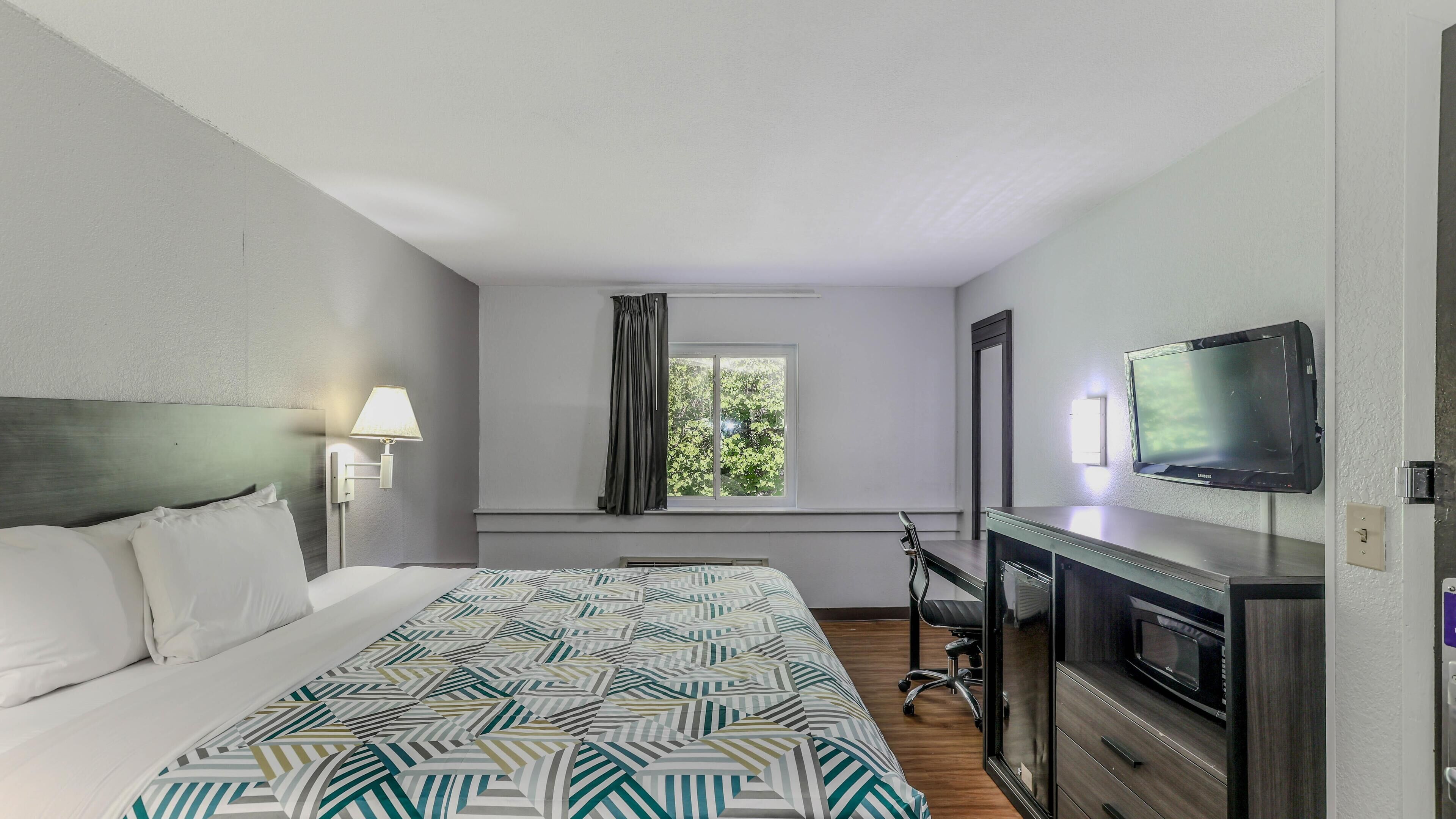 A hotel room with a bed, a TV, a desk, and a window with curtains.