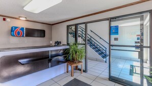 A hotel reception area with a modern counter, a potted plant, and a staircase leading to an upper floor. - Motel 6 Wichita, KS (Wichita)