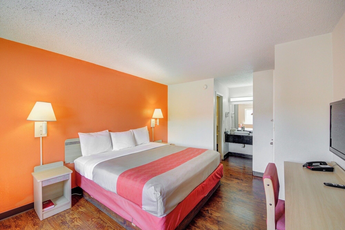 A hotel room with a bed, bedside lamps, a desk with a chair, a television, and a bathroom visible through an open door.