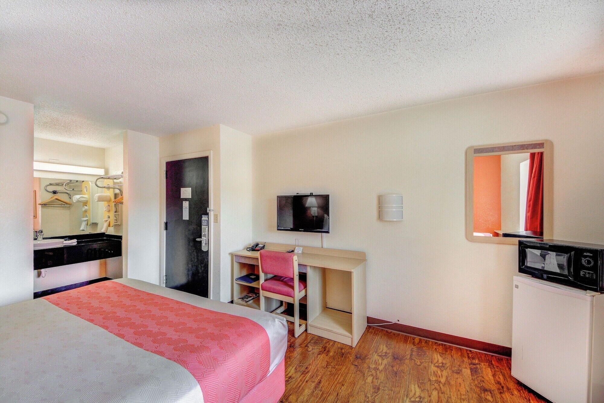 A hotel room with a bed, a desk with a chair, a television, a mirror, and a small refrigerator.
