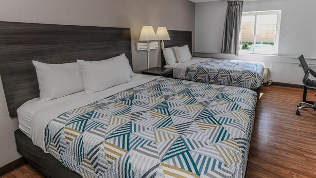 A hotel room with two beds, a dark wood headboard, a patterned bedspread, a nightstand with lamps, a chair, and a window with curtains. - Motel 6 Wichita, KS (Wichita)