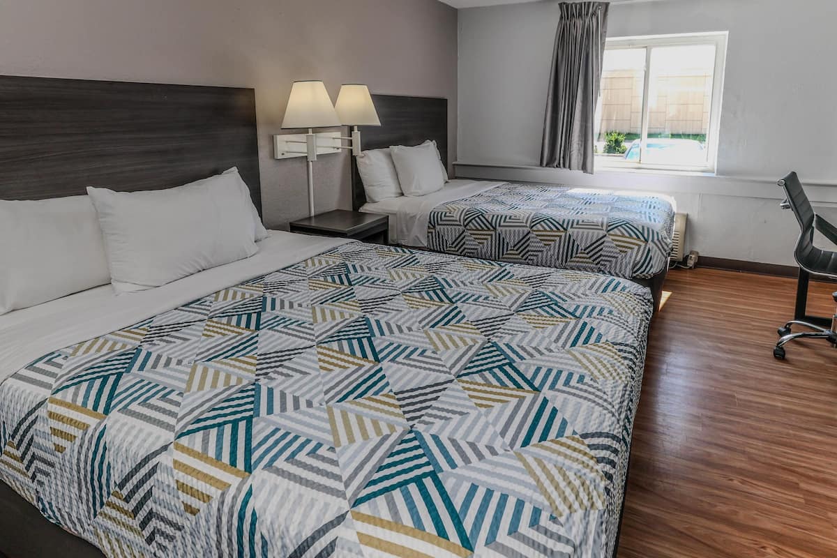 A hotel room with two beds, a dark wood headboard, a patterned bedspread, a nightstand with lamps, a chair, and a window with curtains.