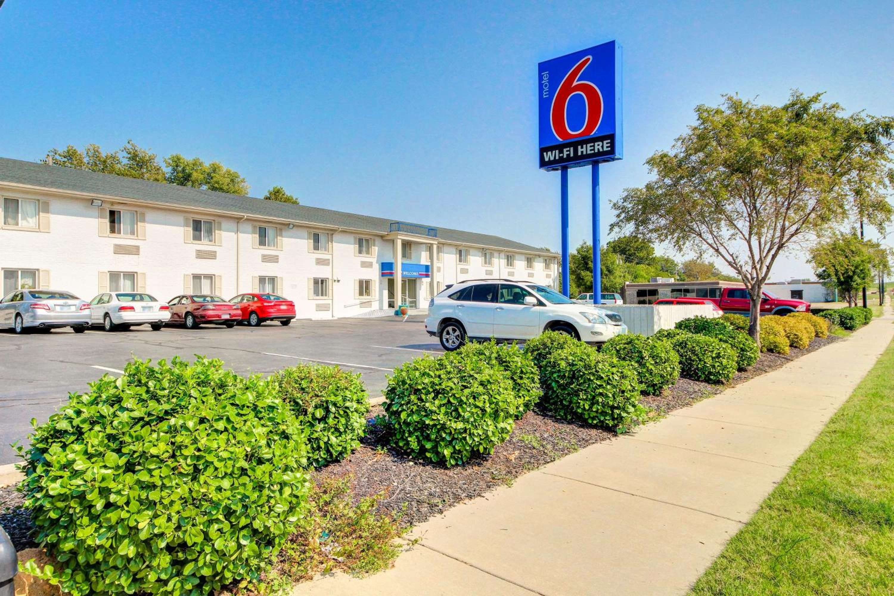 A motel with a large blue sign indicating it is number 6. There are several cars parked in front, and the building has a simple, functional design with a flat roof.