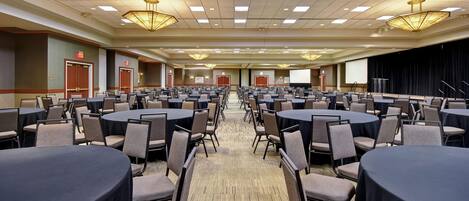 A large conference room with multiple round tables and chairs, a stage with a screen, and a carpeted floor.