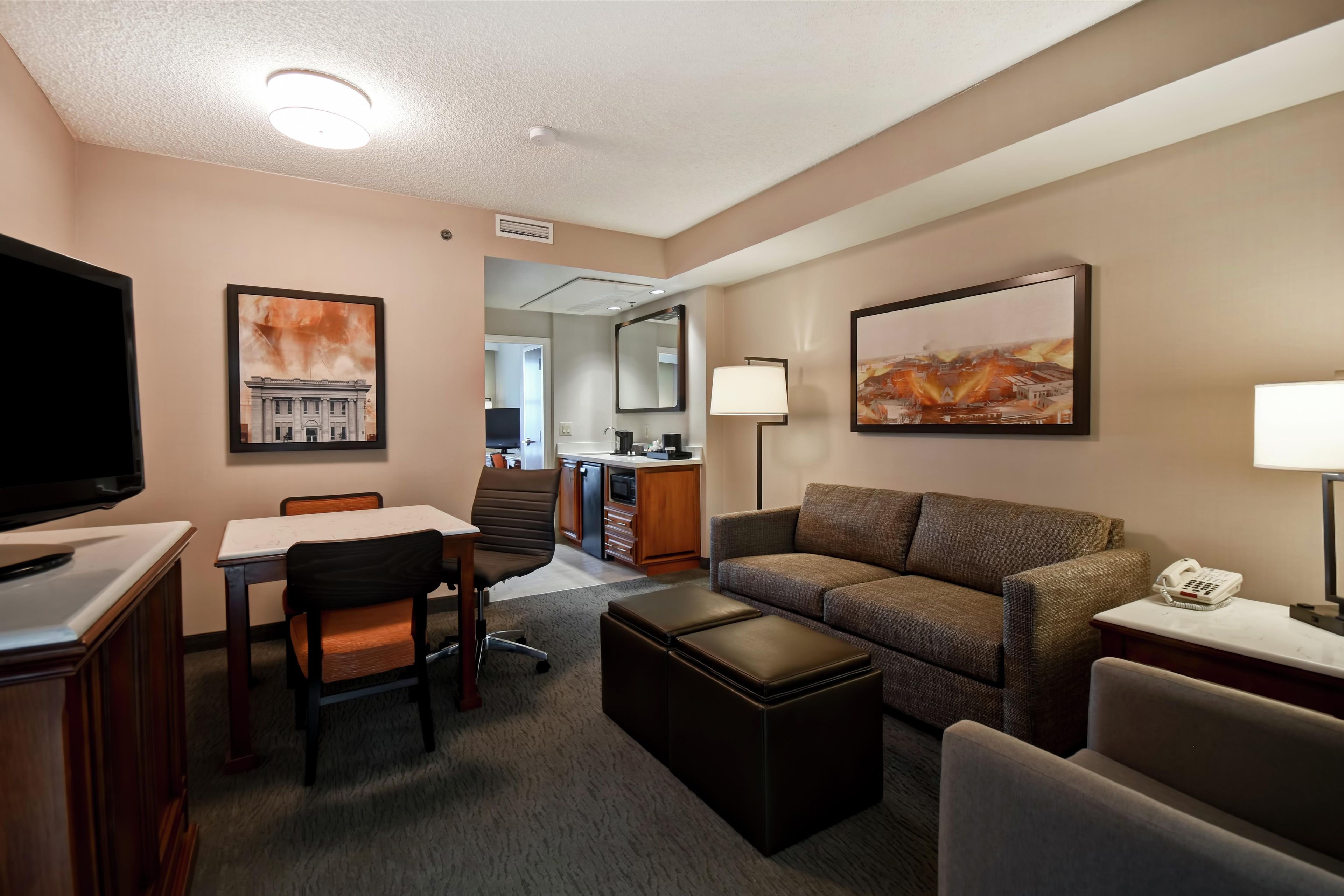 A hotel room with a television, a sofa, a desk with a chair, a coffee table, and artwork on the walls.