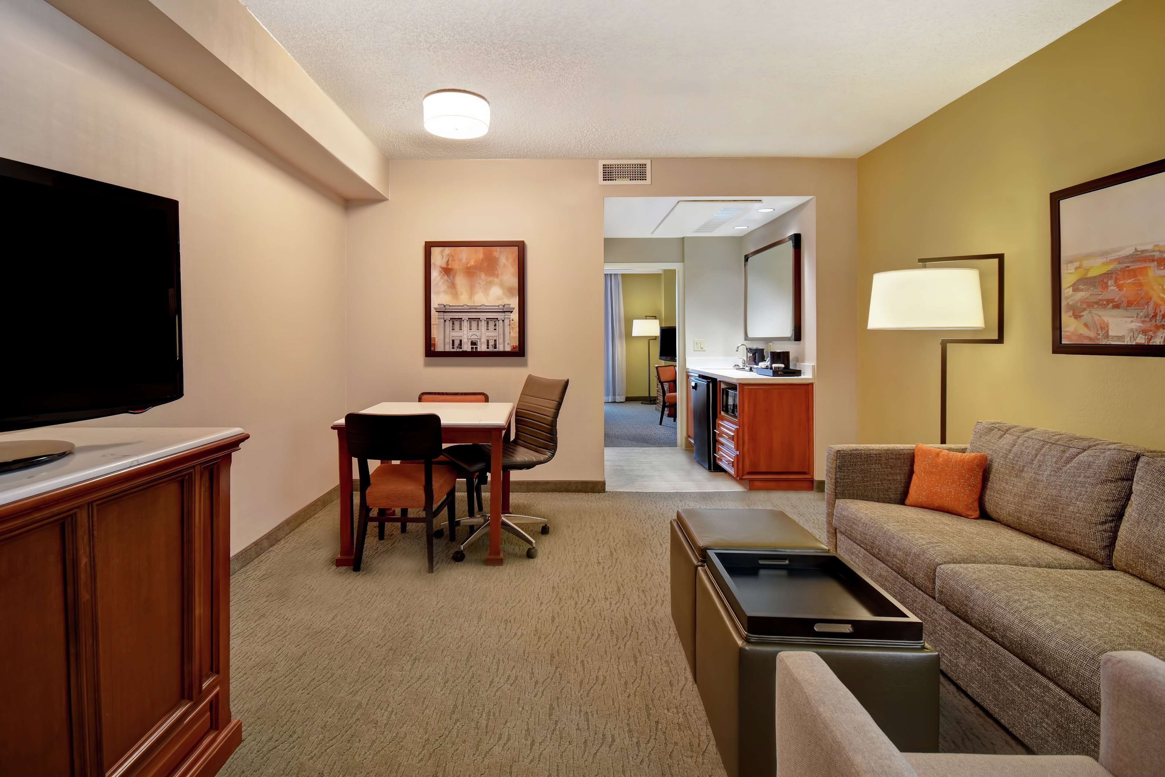 A hotel room with a TV, a dining area with a table and chairs, a sofa, and a desk with a lamp.