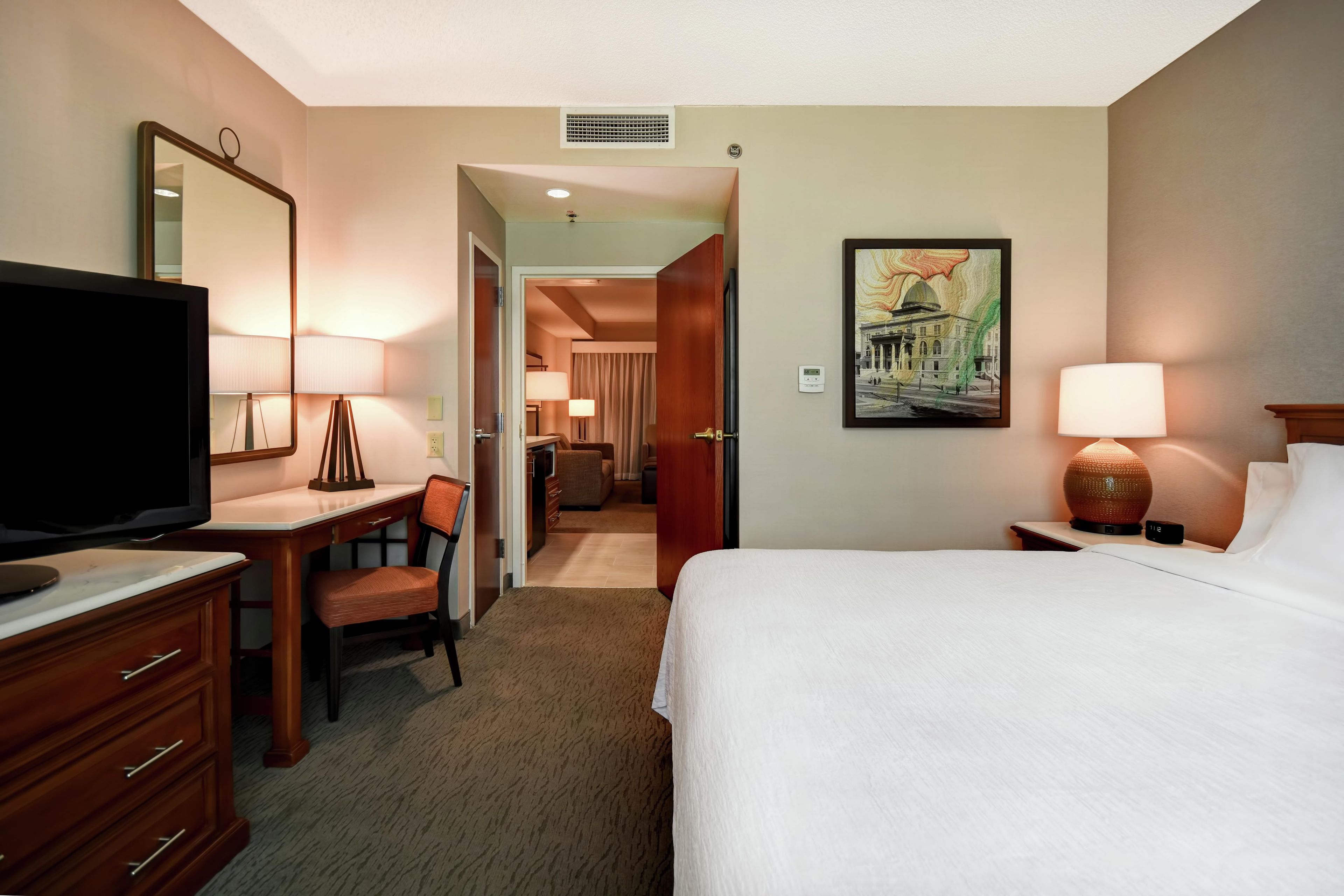 A hotel room with a bed, desk, chair, television, and a painting on the wall.