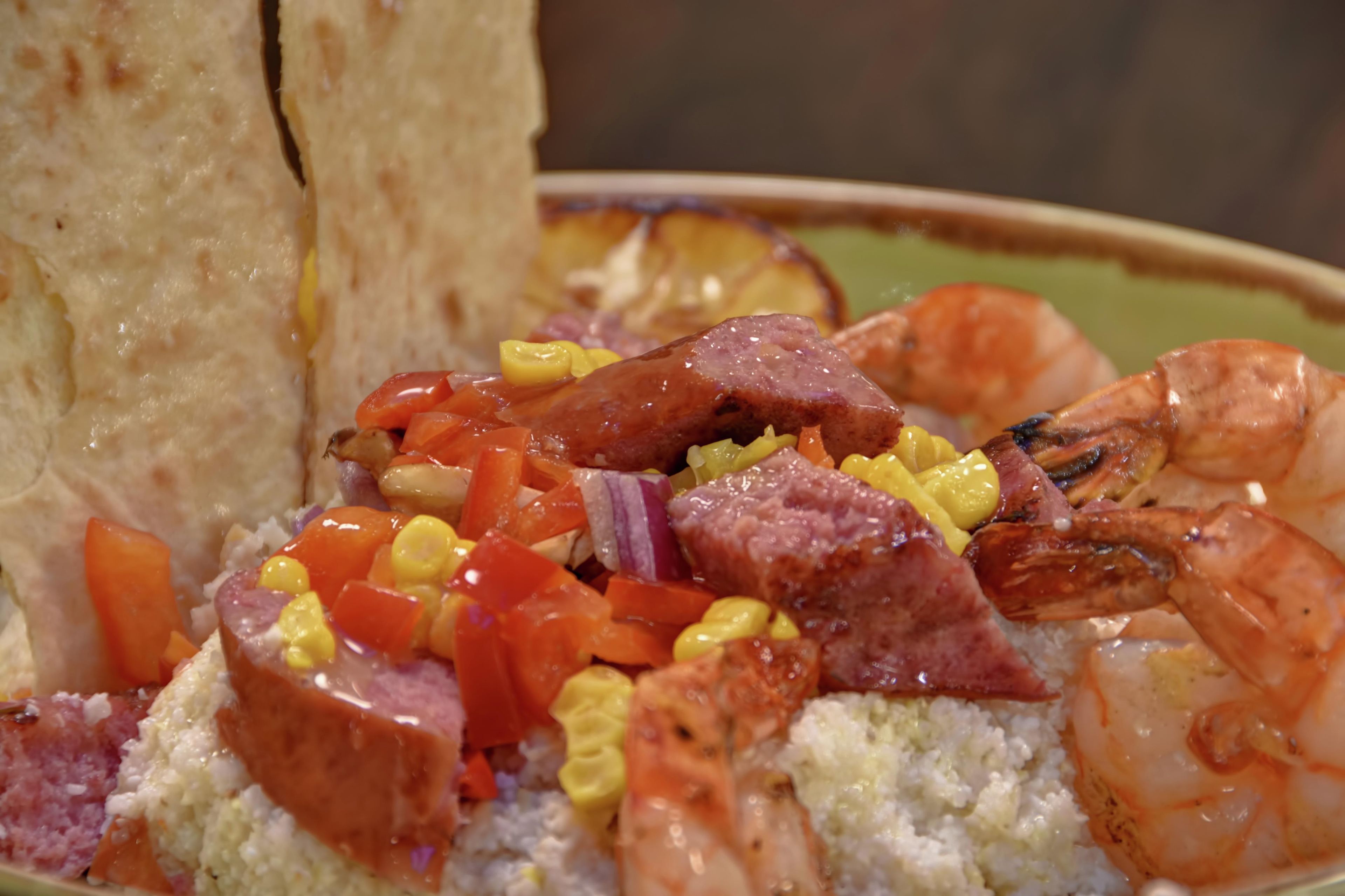 Une assiette de nourriture composée de crevettes, de saucisses et de légumes, le tout servi sur un lit de riz.
