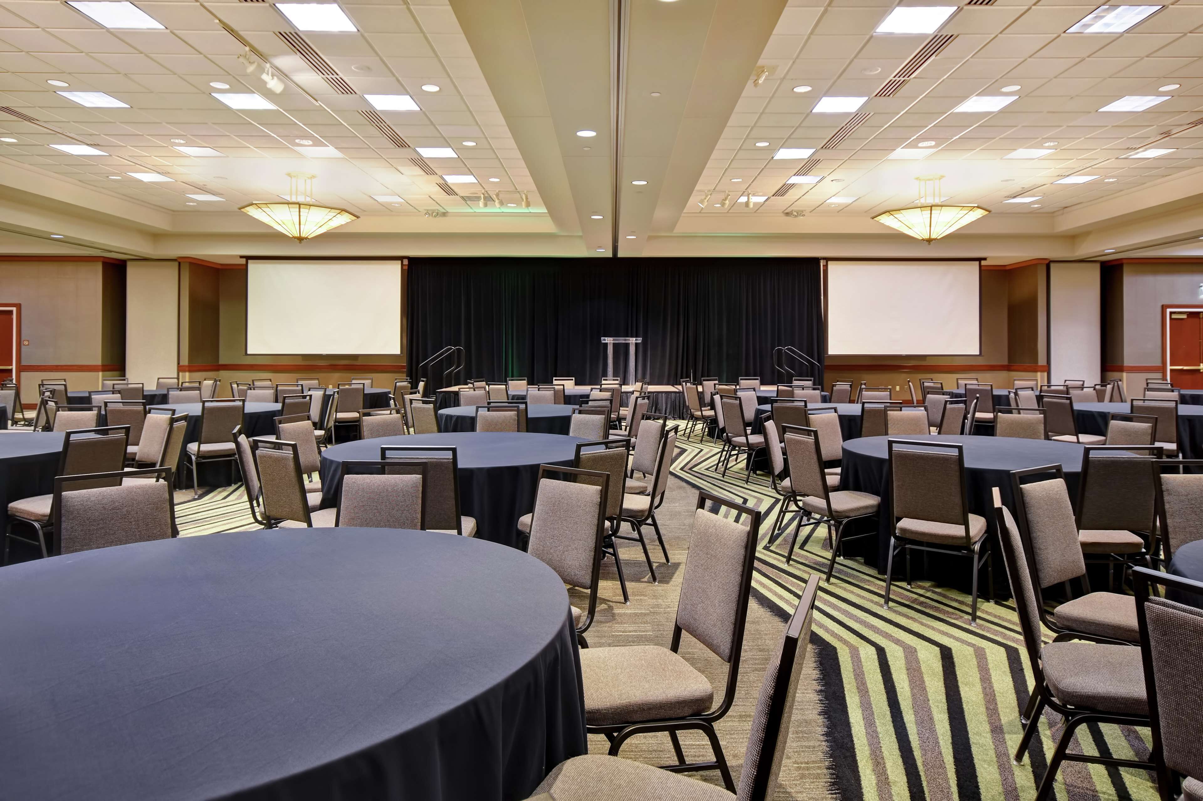 A conference room with multiple tables and chairs, a stage with a screen, and a carpeted floor.