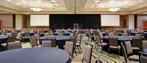 A conference room with multiple tables and chairs, a stage with a screen, and a carpeted floor.