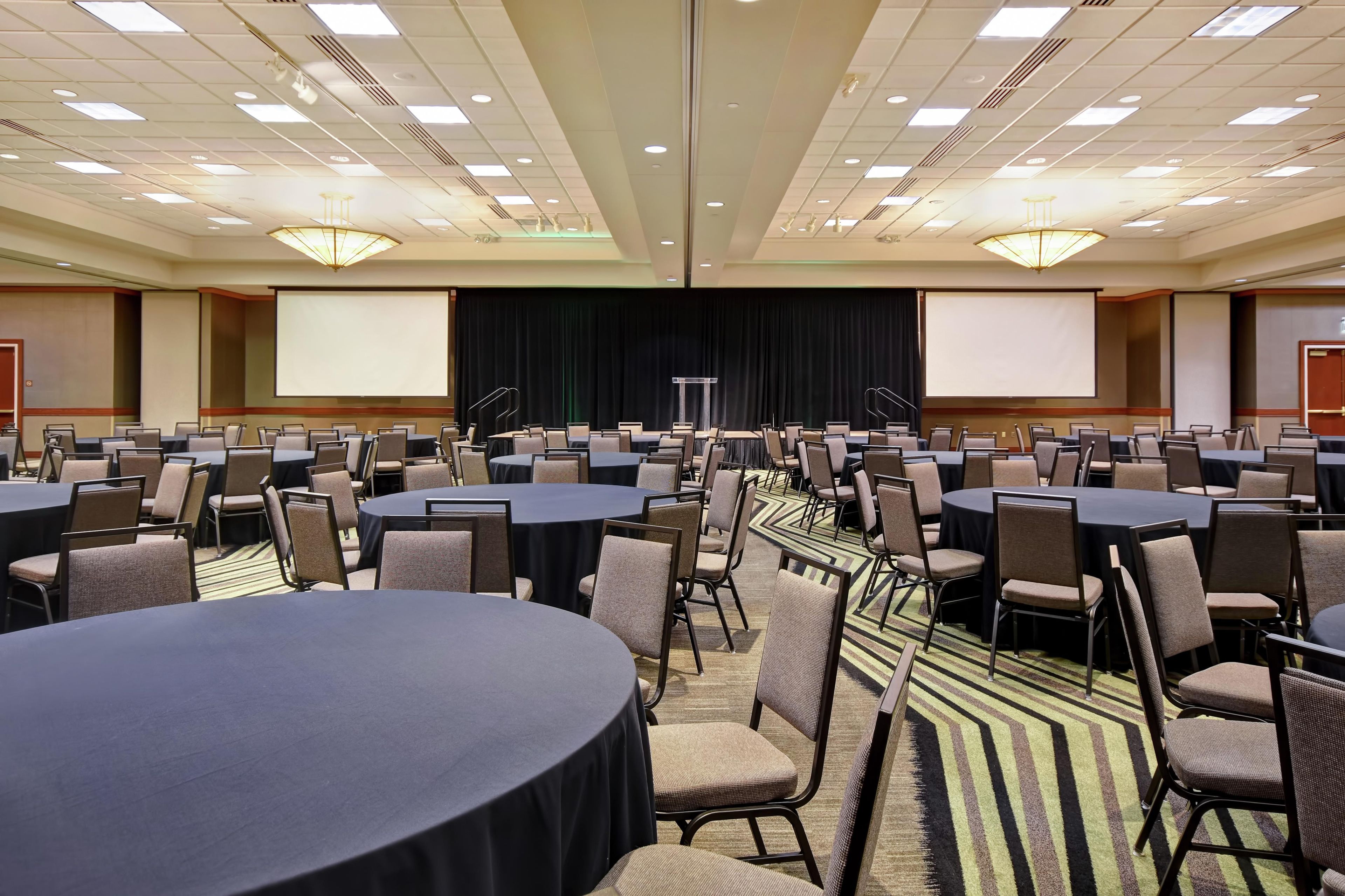Una sala conferenze con diversi tavoli e sedie, un palco con schermo e pavimento rivestito di moquette.