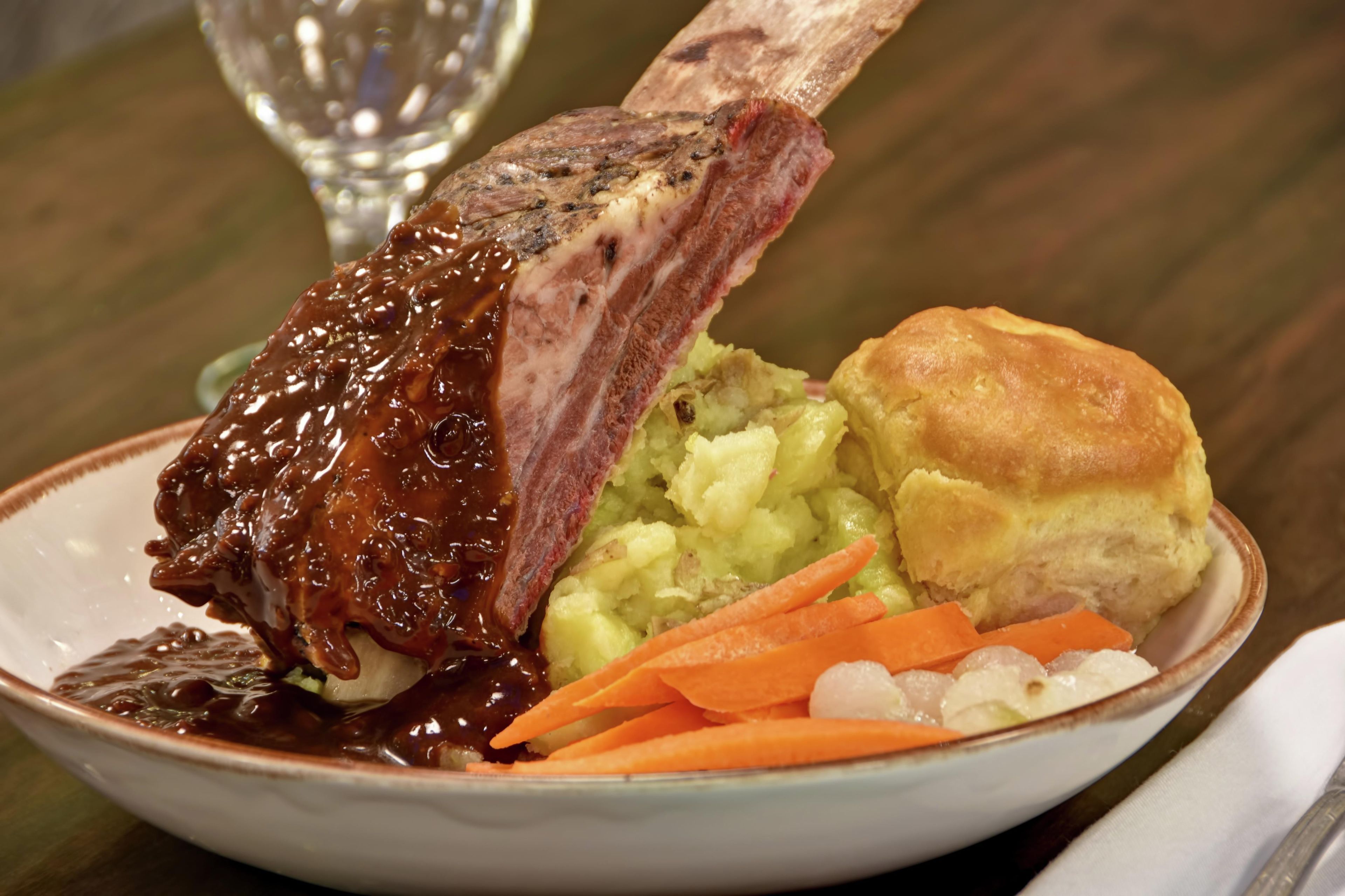 A plate with a bone-in steak, mashed potatoes, carrots, and a biscuit.