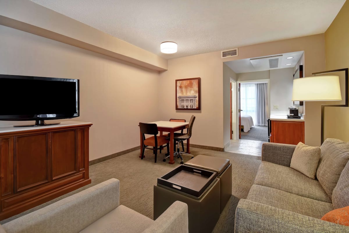 A hotel room with a flat-screen TV on a wooden cabinet, a dining table with chairs, a sofa, a small table with a tray, and a painting on the wall.