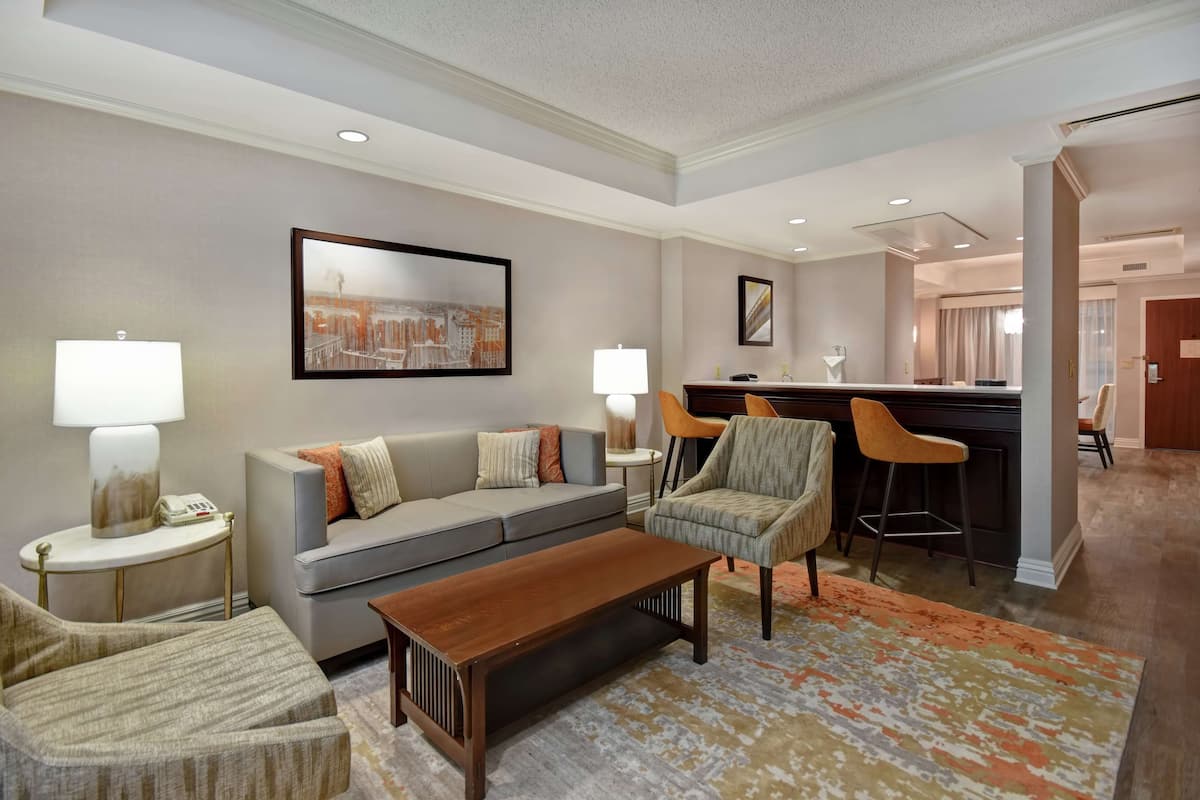 A modern hotel room with a grey sofa, wooden coffee table, and patterned rug. There are two chairs, a bar area, and a framed picture on the wall.