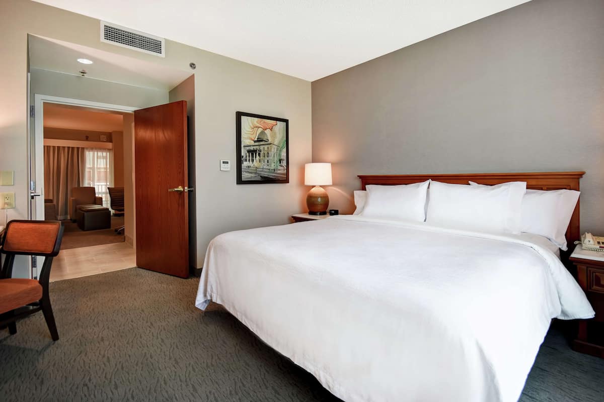 A hotel room with a large bed, a bedside table with a lamp, a chair, and a door leading to another room.