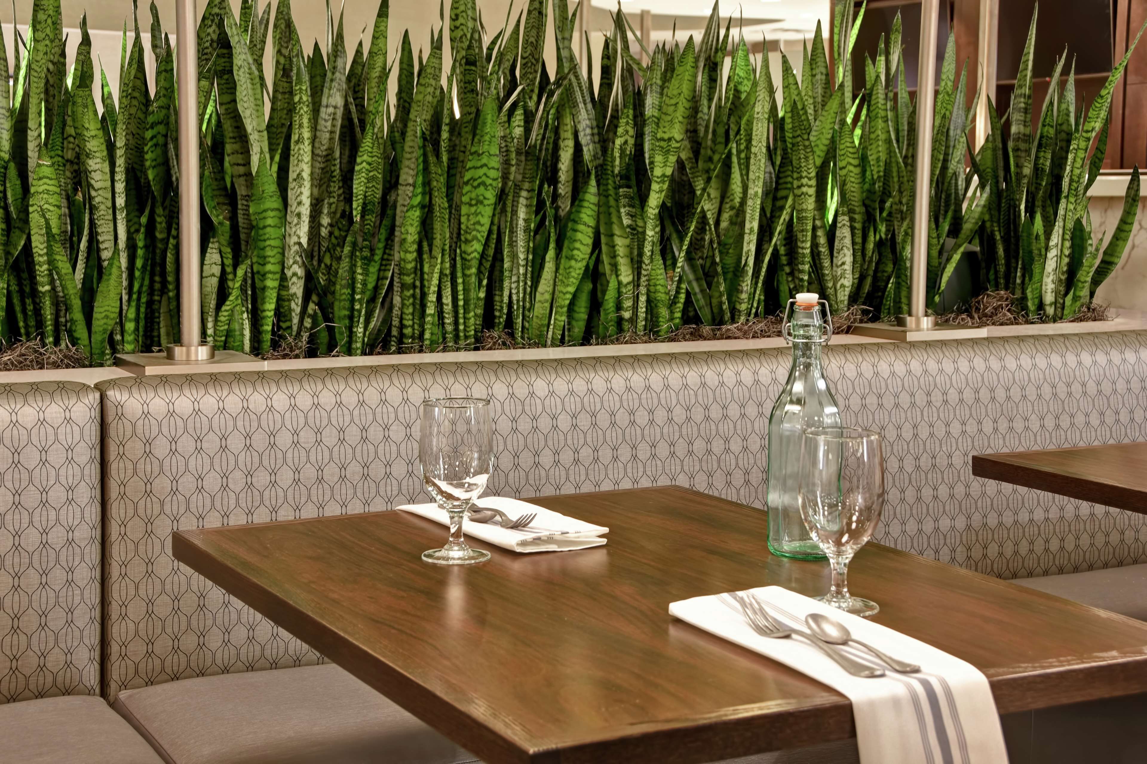 A dining area with a wooden table set for two, a patterned wall, and a row of green plants behind the table.