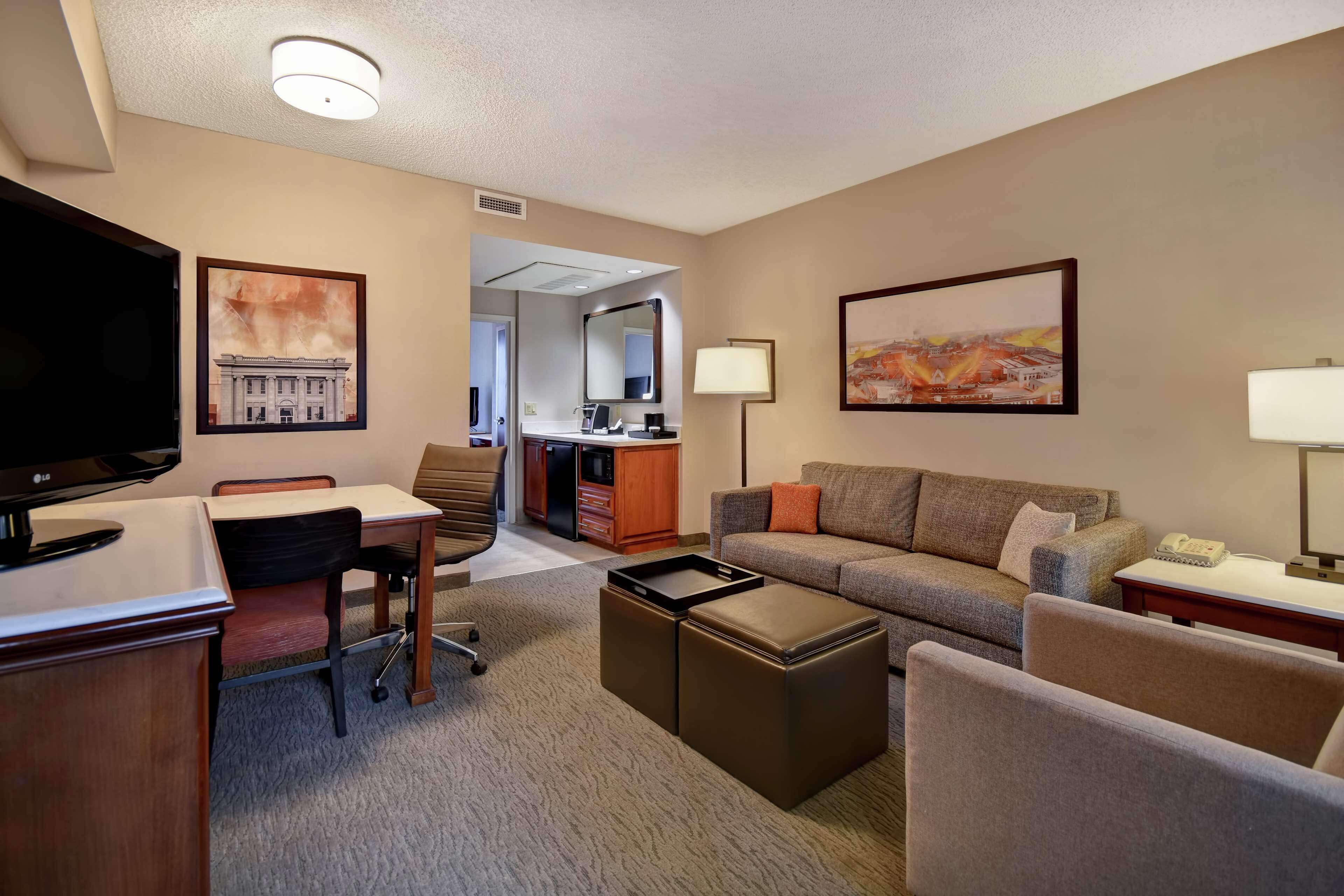A hotel room with a flat-screen TV, a sofa, a desk with a chair, a small table, and a lamp.