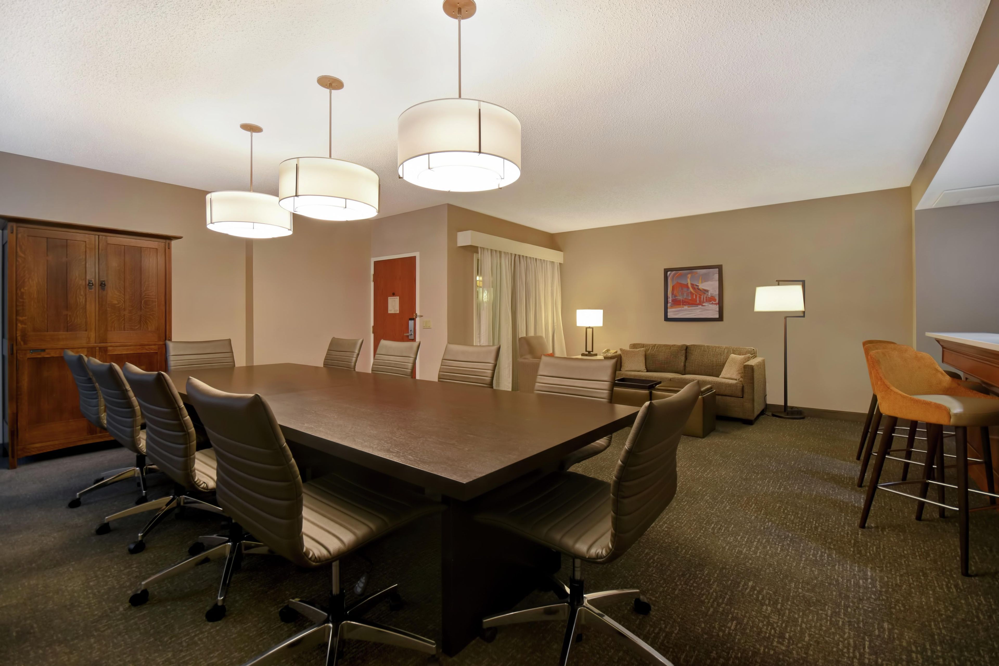 A conference room with a large table, chairs, a sofa, a desk with a lamp, and a painting on the wall.