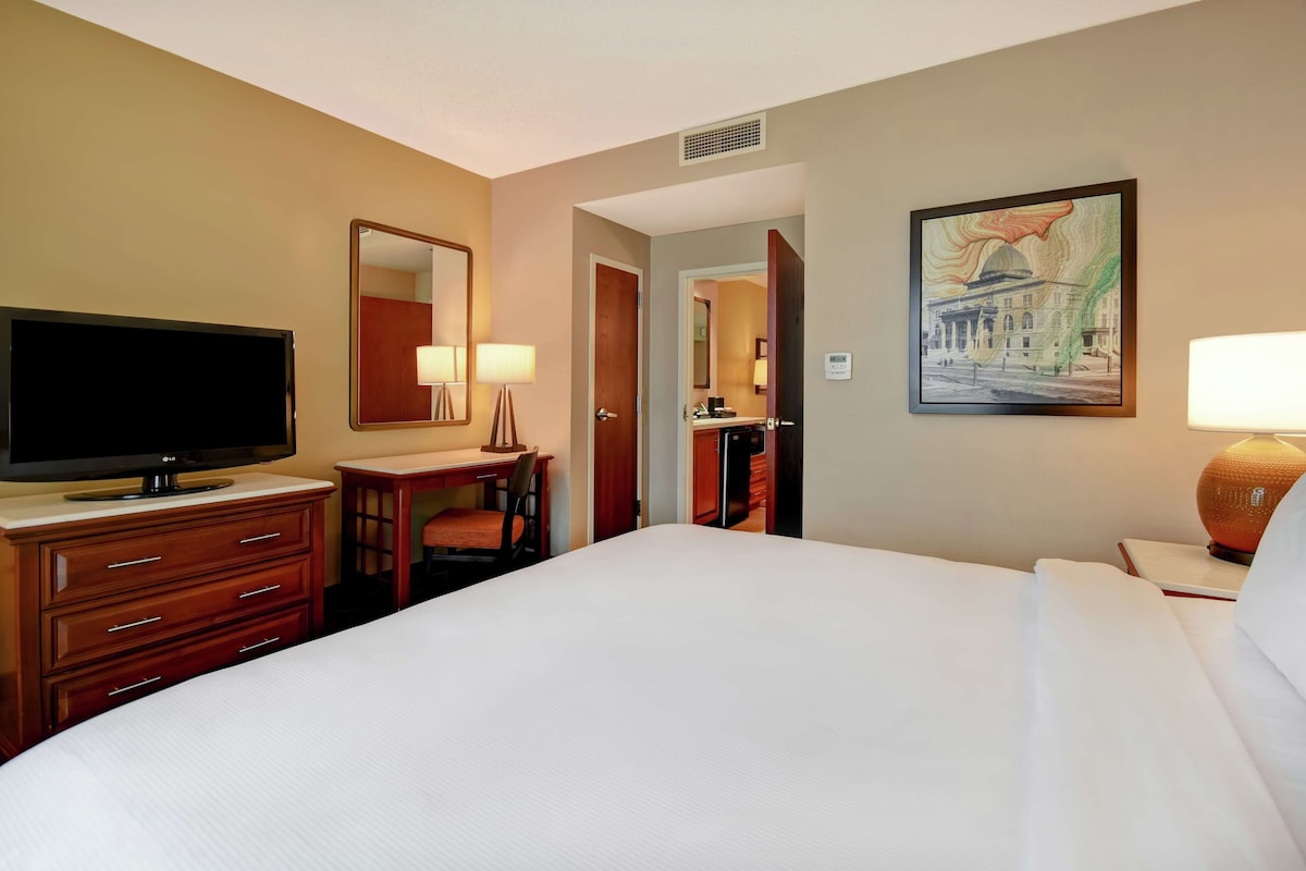 A hotel room with a bed, a desk with a chair, a television, a painting, and a lamp.