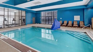 Una piscina interior con agua cristalina, paredes azules y un borde de azulejos. Se observan sillas de descanso y una zona cubierta a la derecha.