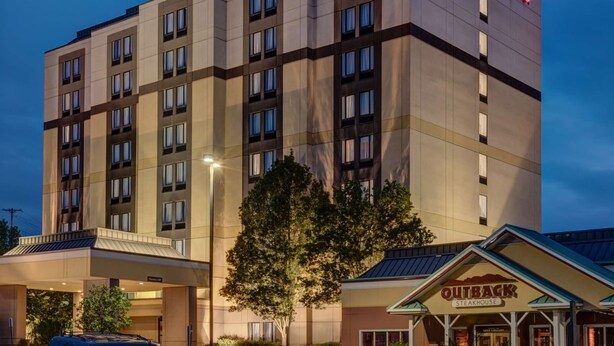 Un hotel Hampton Inn con diseño moderno, que cuenta con un cartel llamativo, una zona exterior cubierta y un restaurante Outback Steakhouse cercano.