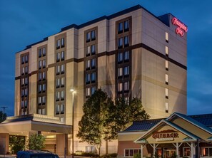 Un hotel Hampton Inn con diseño moderno, que cuenta con un cartel llamativo, una zona exterior cubierta y un restaurante Outback Steakhouse cercano.