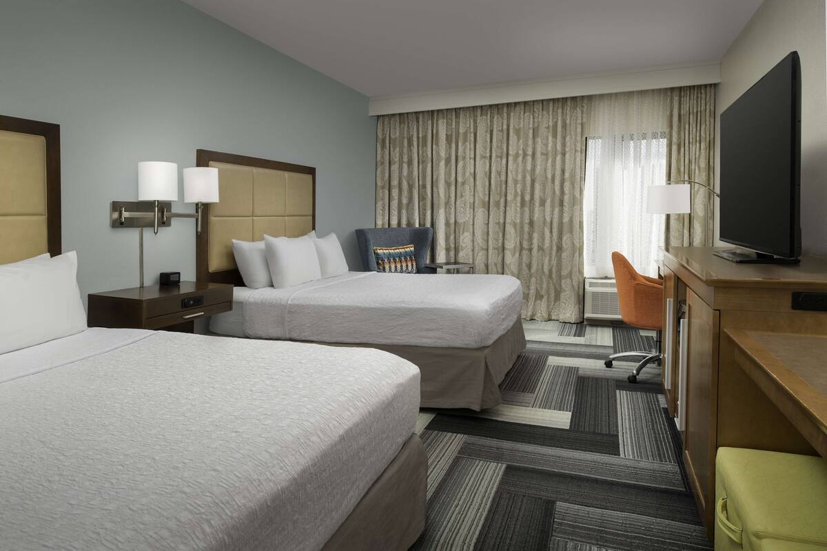 A hotel room with two beds, a desk with a television, a chair, and a large window with curtains.