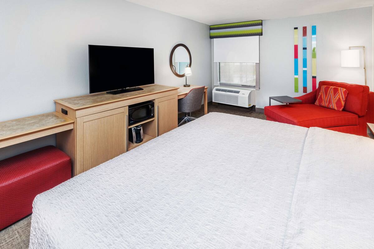Een hotelkamer met een bed, een bureau met een flatscreen-tv, een stoel, een rode bank, een tafeltje en een raam met airconditioning.