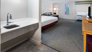 Habitación de hotel con baño moderno, cama con colcha roja, escritorio con computadora y televisor de pared.
