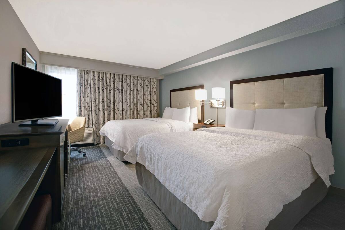 Une chambre d’hôtel avec deux lits, un bureau doté d’un téléviseur à écran plat, une chaise et une grande fenêtre avec des rideaux.