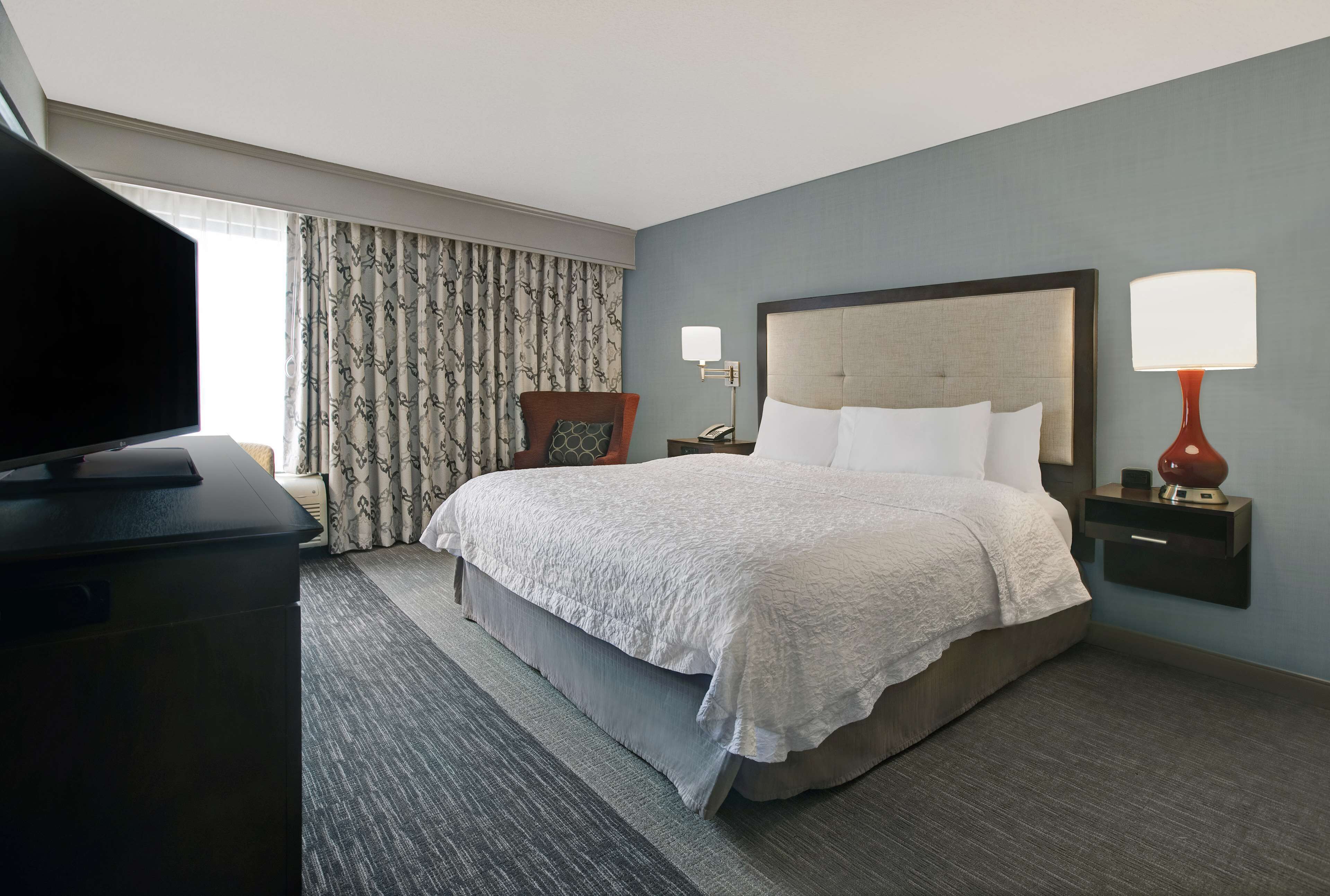 A hotel room with a bed, a television, a chair, a nightstand with a lamp, and a window with curtains.