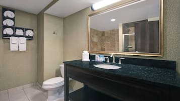 A bathroom with a dark granite countertop, white sink, and a large mirror. There are towels on a shelf and a toilet.