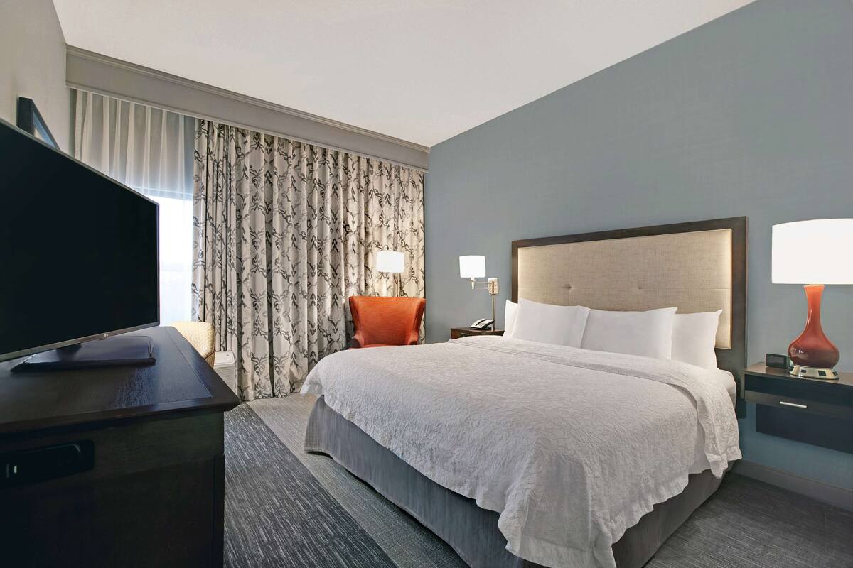 Une chambre d’hôtel avec un grand lit, un téléviseur posé sur une commode, une table de chevet avec une lampe et une fenêtre avec des rideaux.
