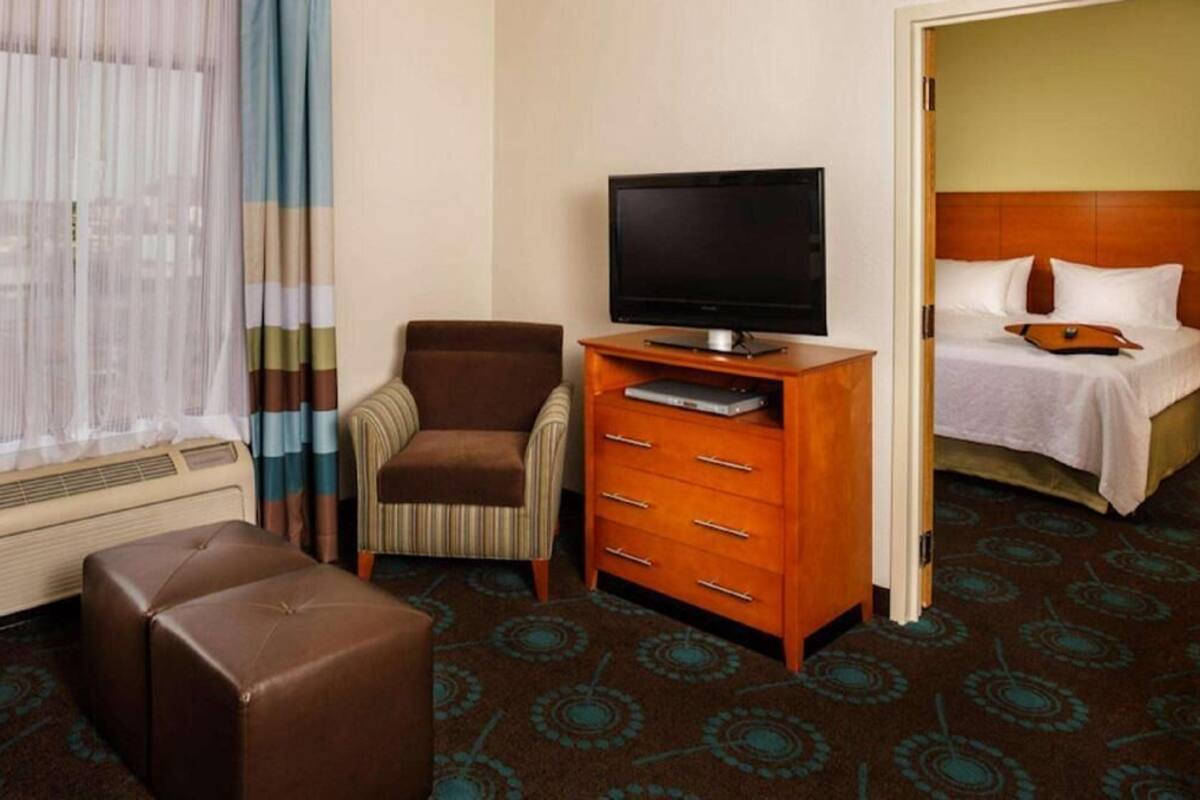 A hotel room with a bed, a television on a wooden stand, a brown armchair, a small ottoman, and a patterned carpet.