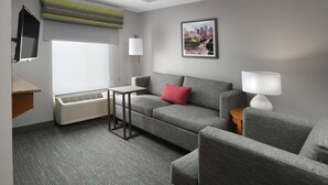 A modern hotel room with a grey sofa, a small table, a red pillow, a cityscape picture on the wall, a flat-screen TV, and a standing lamp. - Hampton Inn & Suites Houston Medical Center NRG Park (Houston)