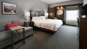A hotel room with a bed, a sofa, a small table, a TV, a mirror, and a window with curtains. - Hampton Inn & Suites Houston Medical Center NRG Park (Houston)