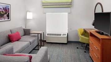 A hotel room with a gray sofa, a small table, a TV on a wooden stand, a yellow chair, and a large window with a view of a cityscape.