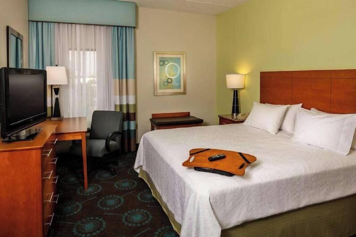 A hotel room with a bed, a desk with a television, a chair, and a nightstand with a lamp.