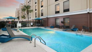 A swimming pool area with lounge chairs and umbrellas, surrounded by a building with multiple windows and a palm tree. - Hampton Inn & Suites Houston Medical Center NRG Park (Houston)