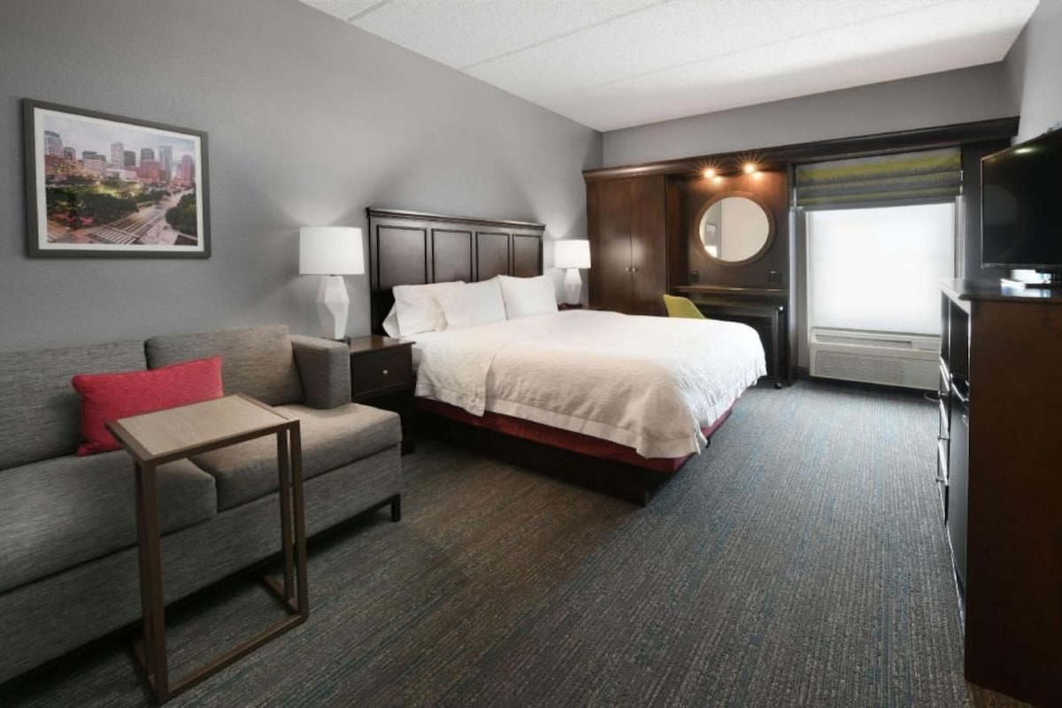 A hotel room with a bed, a sofa, a small table, a TV, and a window with curtains.